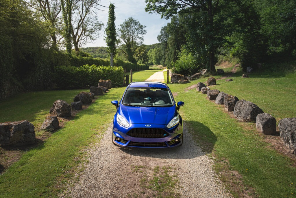 Vue de face d'une Ford Fiesta ST de couleur bleue, garée au centre d'une allée en gravier bordée de verdure et de rochers.