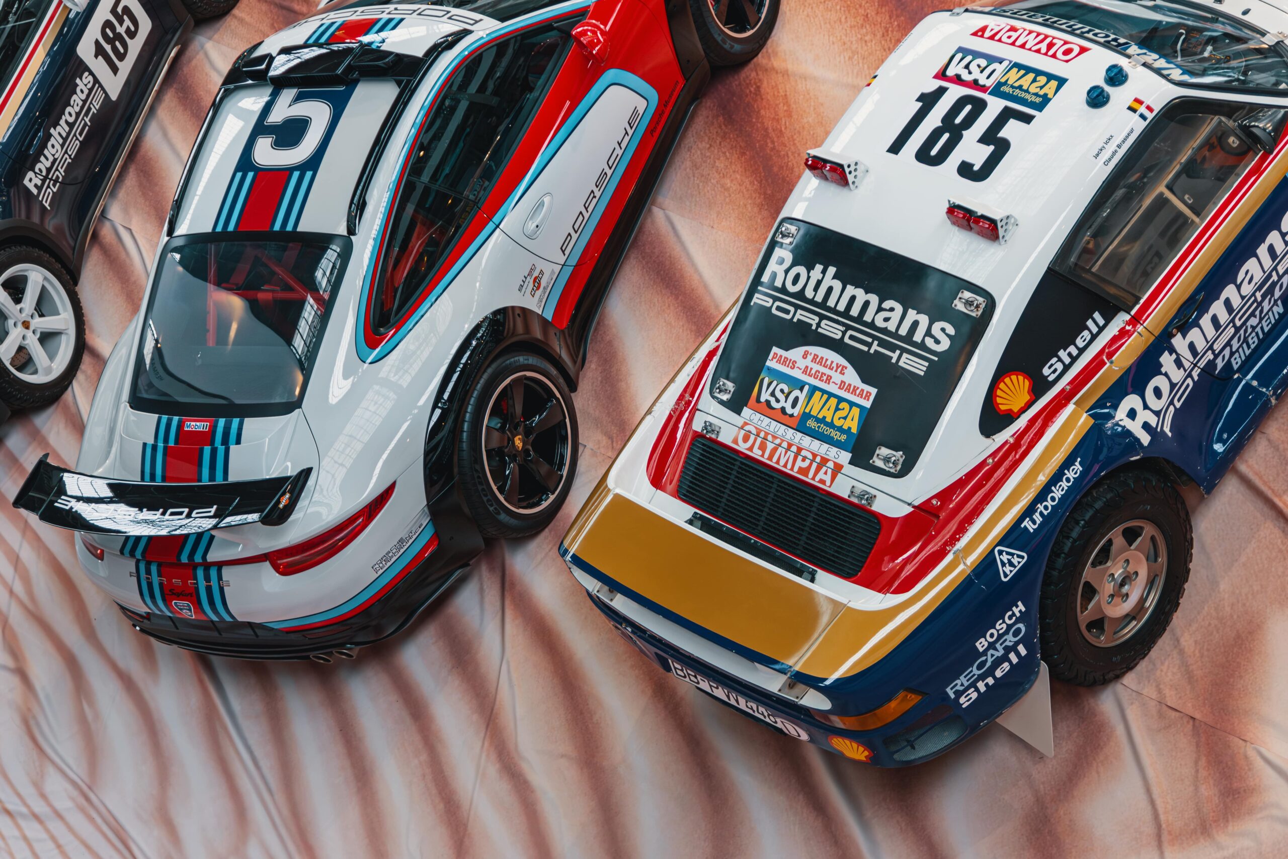 Vue de dessus de deux voitures de course Porsche : à gauche une 911 moderne avec livrée Martini, à droite une 959 classique avec livrée Rothmans du Paris-Dakar.