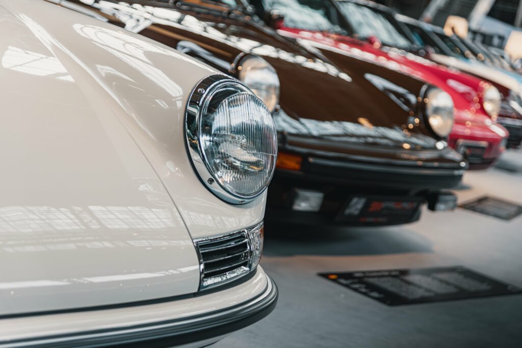 Gros plan sur le phare avant d'une Porsche 911 classique de couleur blanche, avec d'autres modèles anciens alignés derrière elle dans un salon d'exposition.