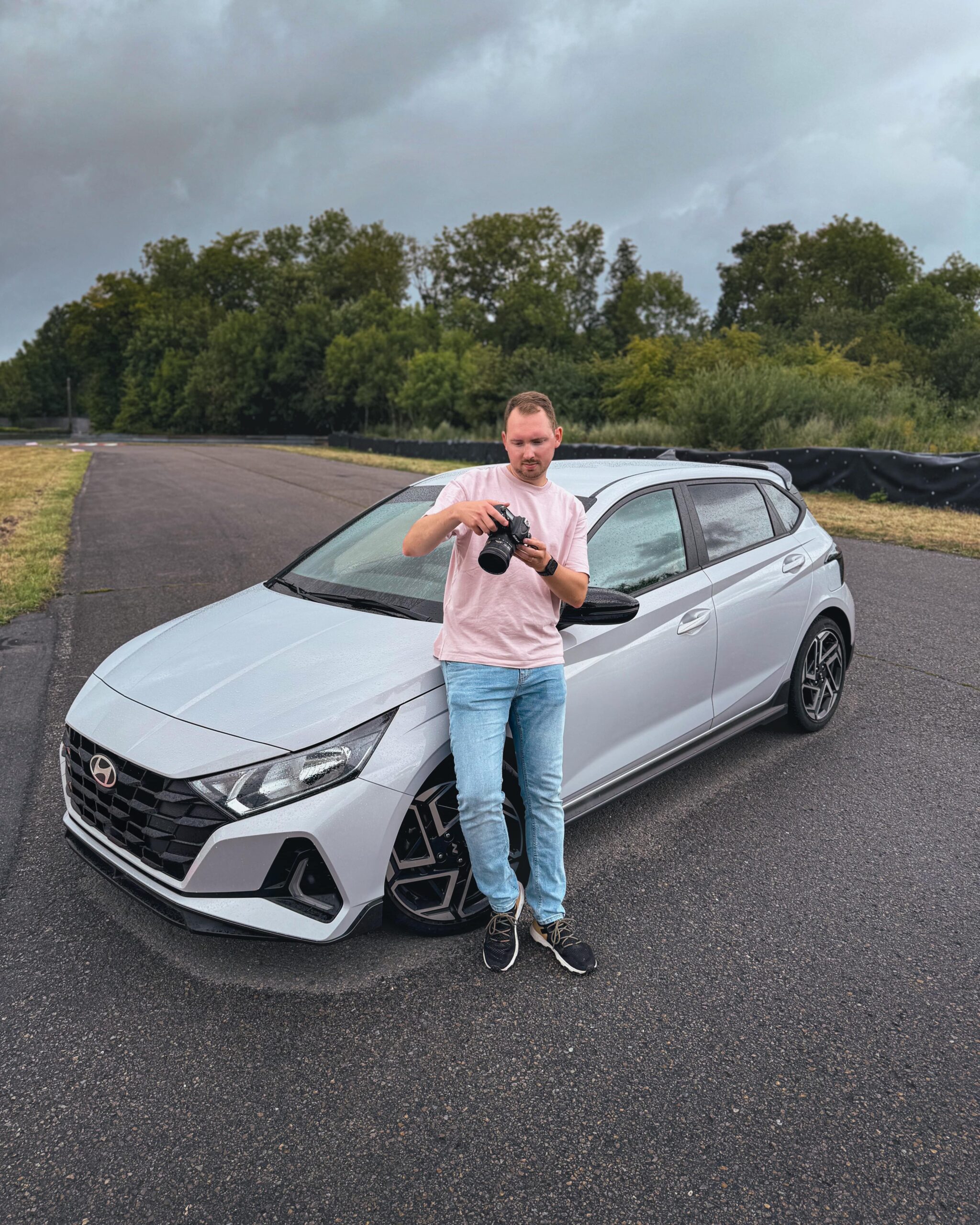 Un homme tenant un appareil photo est appuyé contre le côté d'une Hyundai i20 N-Line grise, garée sur un circuit automobile.
